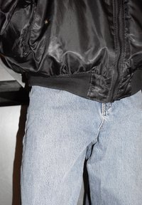 Giacca bomber in raso nero con polsini a costine e cerniera, abbinata a jeans in denim azzurro chiaro con taglio classico e tessuto strutturato.