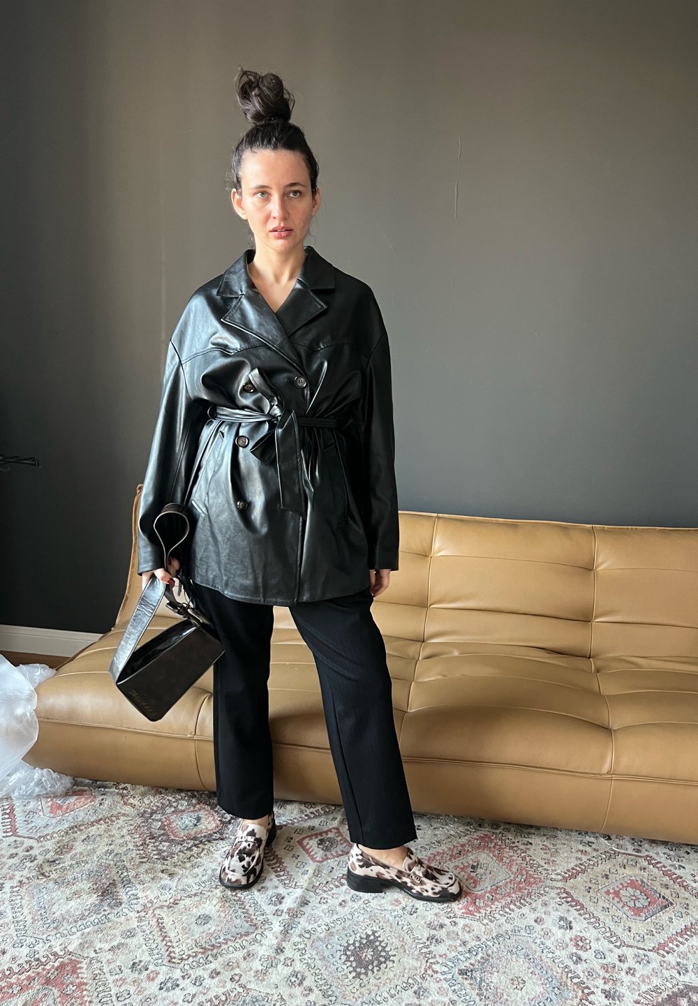 Casaco oversized em pele preta com cinto e botões duplos, combinado com calças pretas e sapatos com padrão. Mala preta elegante.