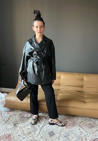 Casaco oversized em pele preta com cinto e botões duplos, combinado com calças pretas e sapatos com padrão. Mala preta elegante.