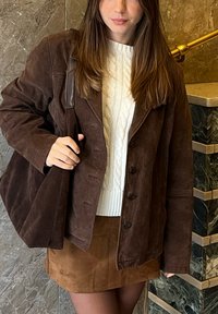 Femme portant un pull torsadé blanc, une veste en suède marron, une jupe marron assortie, et portant un grand sac marron sur l'épaule.