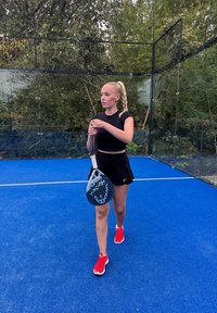 Černý sportovní top, černá sukně, červené boty. Drží šedou pádlo s otvory. Modrý kurt a zelené listoví v pozadí.