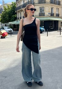 Zwarte asymmetrische tanktop gecombineerd met wijd uitlopende lichtblauwe denim jeans en zwarte sneakers. Vergezeld van oversized zonnebril en een tas met kettinghengsel.