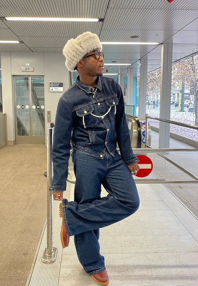 Chaqueta de mezclilla y jeans acampanados en azul oscuro, acentuados con una cadena de perlas y un sombrero de piel gris, situados en una zona de tránsito moderna.