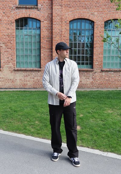 Hombre vistiendo una camisa de manga larga con patrón de cuadros sobre una camiseta gráfica negra, pantalones cargo negros y una gorra negra en un entorno al aire libre.