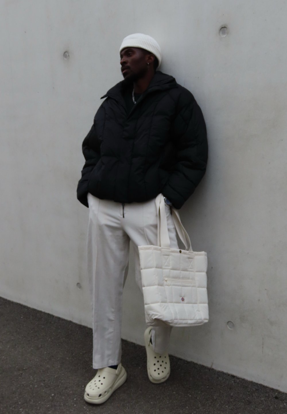 Giacca a vento nera, berretto in maglia bianco, pantaloni beige chiaro, borsa a mano crema e zoccoli bianchi con fori. Superfici testurizzate e design a strati.
