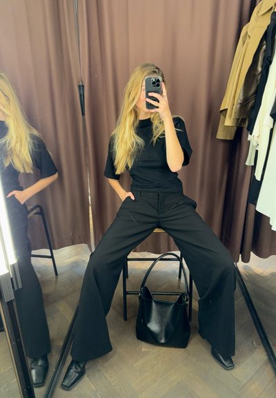 Camiseta negra de manga corta combinada con pantalones negros de pierna ancha y cintura alta. Bolsa negra colocada en el suelo, con espejos reflectantes en el fondo.