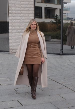 Abrigo beige sobre un vestido de pana marrón, medias marrones y botas marrones hasta la rodilla. Pequeño bolso marrón. Texturas suaves, colores neutros.