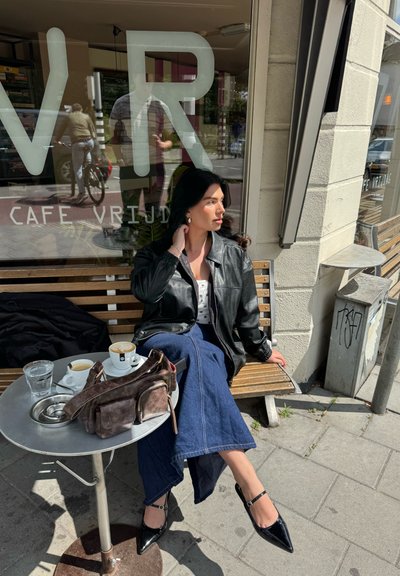 Chaqueta de cuero negra, blusa blanca, falda de mezclilla azul y bailarina negra. Mesa plateada con tazas de café y un bolso marrón.