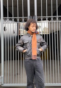 Black leather jacket with chest pockets, an orange-brown scarf with fringes, and dark jeans. Standing against a metal gate backdrop.
