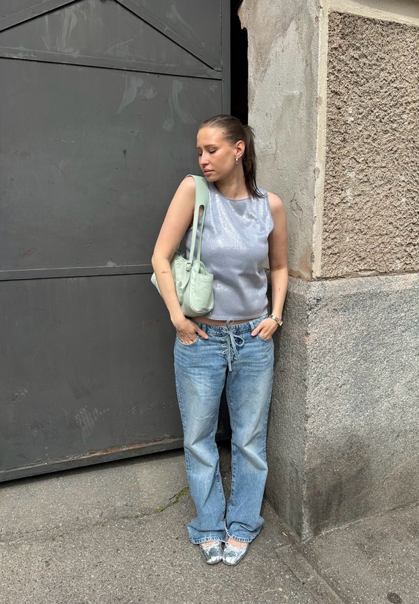 Kvinde med hestehale står mod en gadevæg iført en sølv ærmeløs top, blå jeans, metallic flade sko og bærer en lysegrøn skuldertaske.