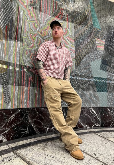 Camisa de cuadros de manga corta en rojo y blanco, pantalones cargo beige con cremalleras, y botas marrones; de pie contra una pared de mosaico colorido.
