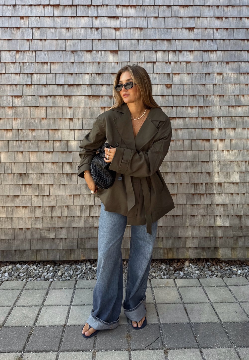 Blazer oversize verde oliva con ampie revers, vita legata, abbinato a jeans larghi di un leggero blu e una pochette nera intrecciata. Occhiali da sole moderni completano il look.