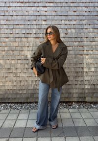 Blazer oversize verde oliva con ampie revers, vita legata, abbinato a jeans larghi di un leggero blu e una pochette nera intrecciata. Occhiali da sole moderni completano il look.