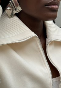 Vit zip-jacka med en texturerad krage, tillverkad av mjukt tyg. Har silverdetaljer och en slät yta. Närbild på halslinningen.