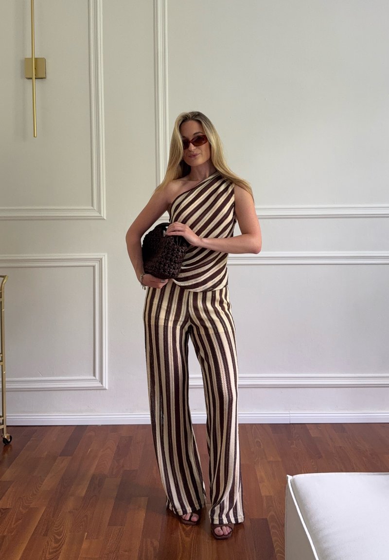 Femme portant un haut à rayures verticales marron et crème à une épaule et un pantalon large assorti, tenant un sac à main tissé foncé à l'intérieur.