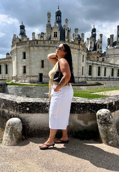 Falda blanca hasta la rodilla, parte superior beige y bolso negro. El fondo presenta una arquitectura de castillo ornamental con oscuros pináculos contra un cielo nublado.