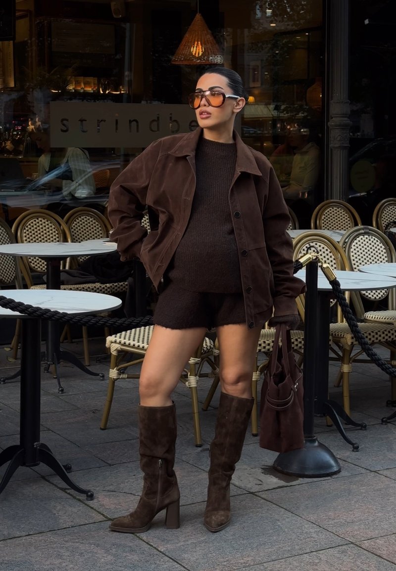 Woman wearing brown jacket, knit top, shorts, tall boots, and sunglasses stands outside café with wicker chairs and marble tables.