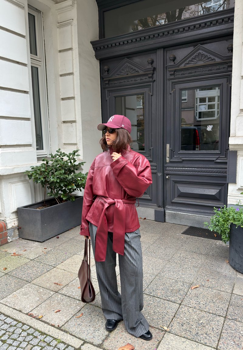 Personne portant une veste en cuir marron rougeâtre et une casquette, un pantalon gris rayé, tenant un sac marron, debout sur un trottoir devant de grandes portes en bois sombre.