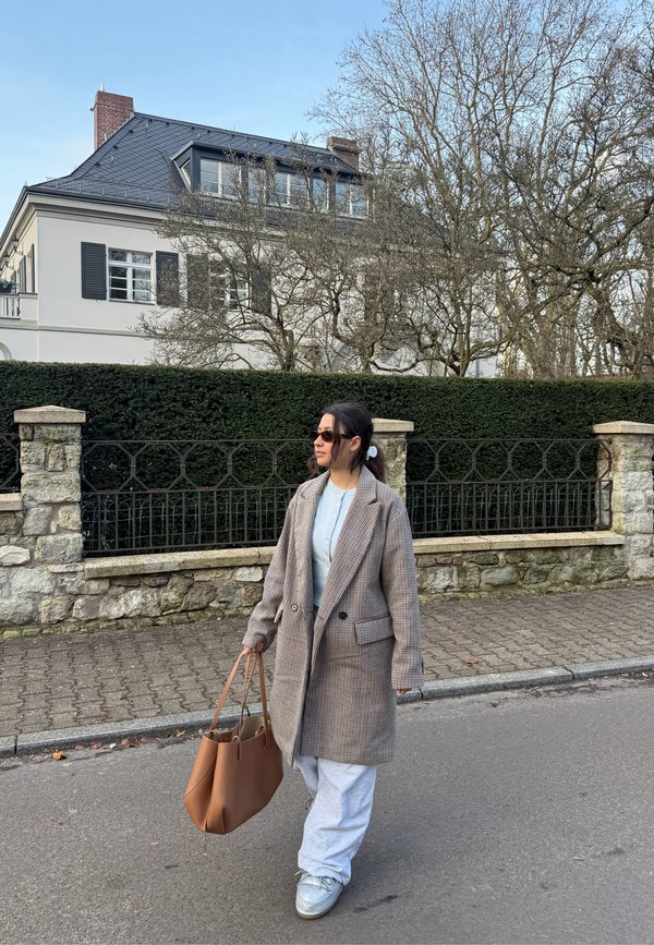 Manteau marron à carreaux à double boutonnage, haut bleu clair, pantalon de survêtement gris, portant un sac beige et des baskets claires, marchant dans une rue.