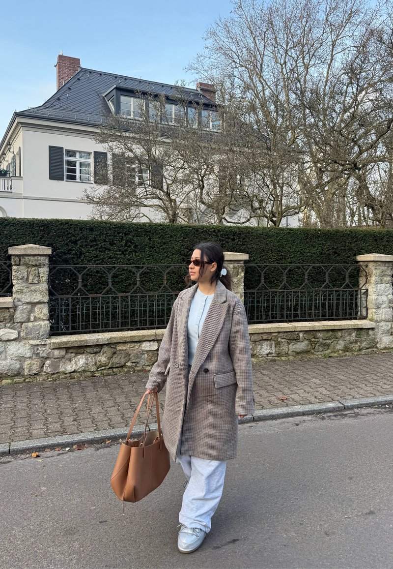Manteau marron à carreaux croisé, haut bleu clair, pantalon de survêtement gris, portant un sac beige et des baskets claires, marchant dans une rue.