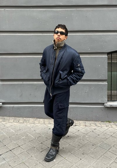 Hombre con chaqueta bomber azul oscuro, pantalones cargo, botas negras y gafas de sol, de pie con las manos en los bolsillos sobre una acera de baldosas.