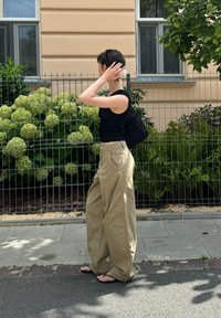 Mustat lyhyet neulepaidat, löysät beige-karkkipuntit ja flip-flopit. Hiukset lyhyet, musta kuvioitu olkalaukku. Taustalla vihreitä pensaita.