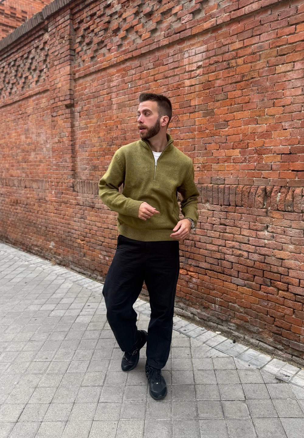 Homme en pull vert olive et pantalon noir marchant sur un trottoir carrelé à côté d'un grand mur de briques rouges, regardant vers la gauche.