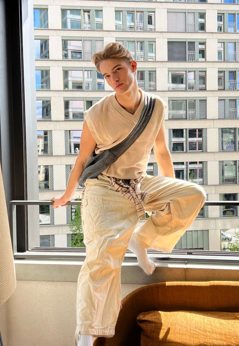 Young person in beige sleeveless sweater and cargo pants sits on windowsill inside apartment with tall building visible outside.