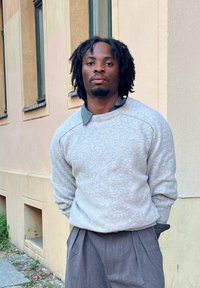 Svetlosivý sveter s textúrovanou látkou, s pásikovým golierom a tmavou pruhovanou košeľou pod ním, kombinovaný s tmavosivými plisovanými nohavicami.