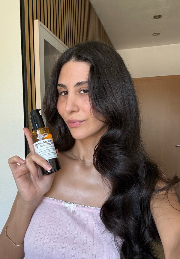 Femme aux longs cheveux ondulés foncés tenant une bouteille de L'Oréal Absolut Repair Molecular Oil, à l'intérieur avec des murs en bois et blancs.
