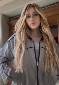 Jachetă cu fermoar de culoare gri deschis, cu accente negre, manșete strânse și design sportiv. Textură netedă, purtată peste un top gri. Păr lung, ondulat.