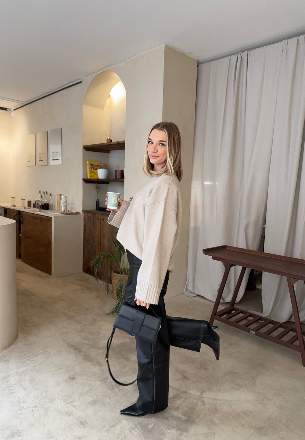 Γυναίκα με μπεζ πουλόβερ και μαύρο δερμάτινο παντελόνι που κρατά κούπα καφέ και μαύρη τσάντα, στέκεται μέσα σε εσωτερικό χώρο με το ένα πόδι σηκωμέν ο και χαμογελάει.