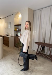 Γυναίκα με μπεζ πουλόβερ και μαύρο δερμάτινο παντελόνι που κρατά κούπα καφέ και μαύρη τσάντα, στέκεται μέσα σε εσωτερικό χώρο με το ένα πόδι σηκωμένο και χαμογελάει.