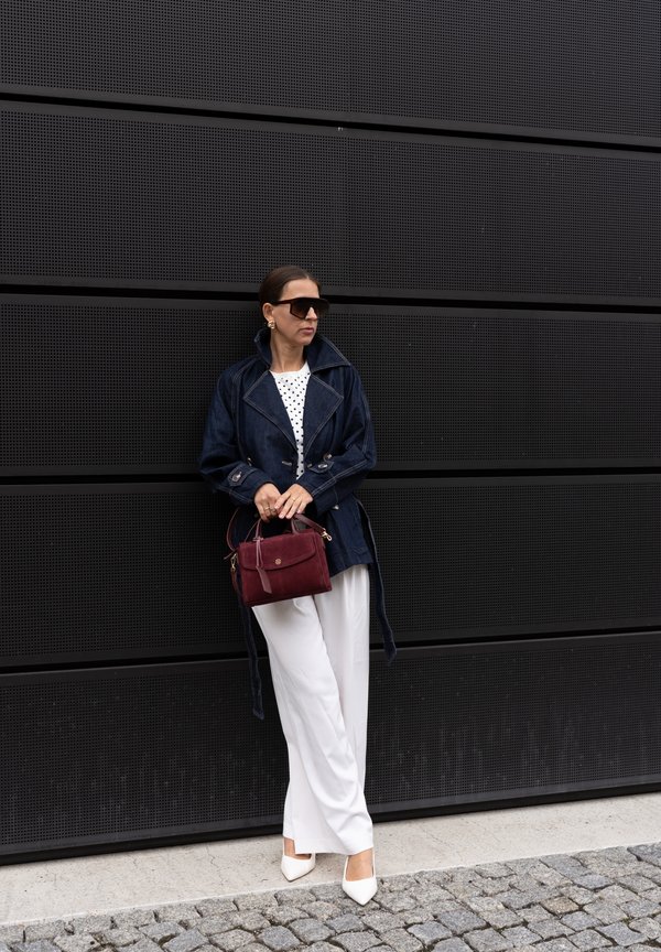Femme en veste sombre, pantalon blanc et lunettes de soleil tenant un sac à main bordeaux, debout contre un mur métallique noir perforé.