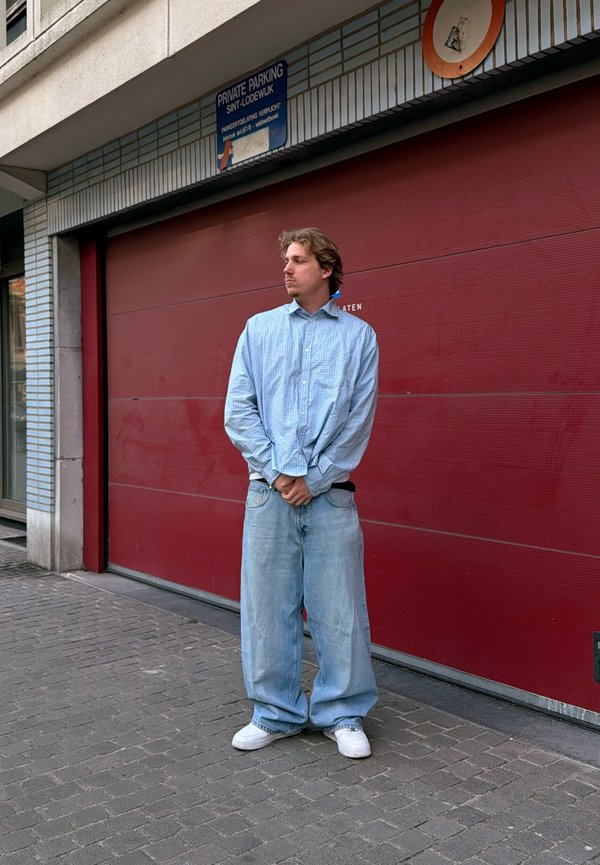 Ung person står på brosteinsbelagt fortau iført løse blå jeans, lyseblå rutete skjorte og hvite joggesko foran en lukket rød garasjeport.