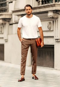 Pantaloni in lino marroni, t-shirt bianca a girocollo e mocassini marroni. Tiene una piccola borsa in pelle marrone. Linee semplici e pulite, stile casual.
