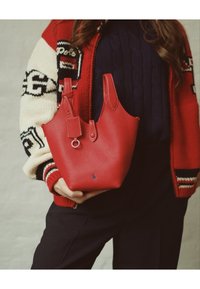 Osoba u crvenoj i bijeloj pletenoj jakni drži teksturiranu crvenu kožnu torbu ispred tamnoplave veste i crnih hlača.