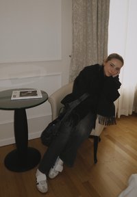 Châle noir à franges sur un haut foncé, jean gris à jambes larges, baskets blanches à Velcro, assis sur une chaise crème à côté d'une table avec un dessus en verre.