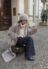 Manteau en laine beige sur un pull blanc, pantalon large à carreaux, et sac à main foncé avec des accents dorés. Chapeau en fourrure grise. Fond pavé.