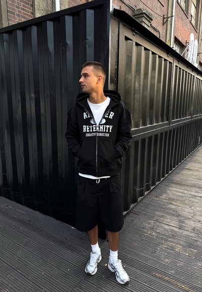 Sudadera negra con cremallera y estampado blanco, combinada con pantalones cortos negros y zapatillas grises. El fondo presenta una pared negra texturizada.