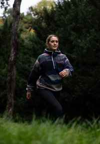 Fleeceová mikina v duhových vlnách modré, fialové a růžové barvy s vysokým límcem. V kombinaci s černými legínami, umístěná v zeleném venkovním prostředí.