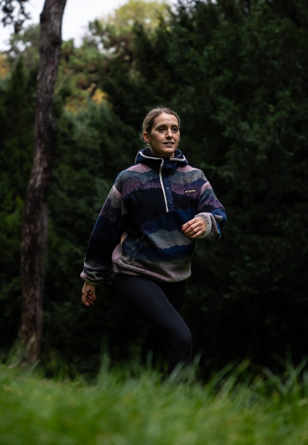 Sudadera de forro polar en ondas multicolores de azul, morado y rosa con un cuello alto. Combinada con leggins negros, ambientada en un entorno natural verde.