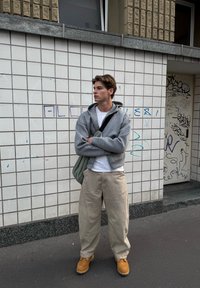 Grauer Reißverschluss-Hoodie über einem weißen T-Shirt, beigen weit geschnittenen Hosen und braunen Arbeitsstiefeln. Eine schlanke Tasche in hellgrün hängt über einer Schulter.