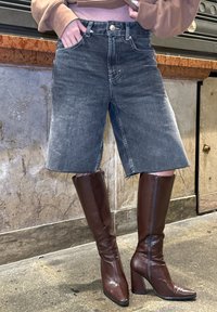 Denim-Bermuda-Shorts in verblasstem Grau mit fransigem Saum, kombiniert mit hohen, polierten braunen Stiefeln mit Absatz. Fokus auf modernem Schnitt und Texturen.
