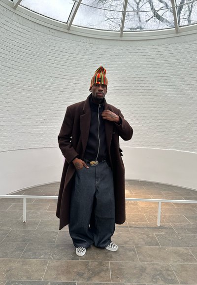 Hombre con abrigo largo marrón oscuro, gorro de punto de rayas coloridas, jeans oscuros y sueltos, y zapatos de cuadros sin cordones, de pie en una habitación circular de ladrillos blancos.