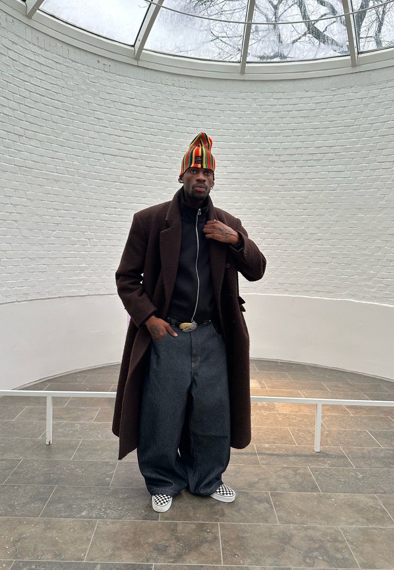 Homme portant un long manteau marron foncé, un bonnet tricoté rayé coloré, un jean ample foncé et des chaussures à enfiler à carreaux, debout dans une pièce circulaire en briques blanches.
