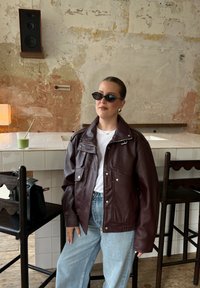 Donna con giacca di pelle marrone, camicia bianca, jeans blu e occhiali da sole neri sta in piedi accanto a un bar piastrellato con sedie in legno e un altoparlante a muro a vista.