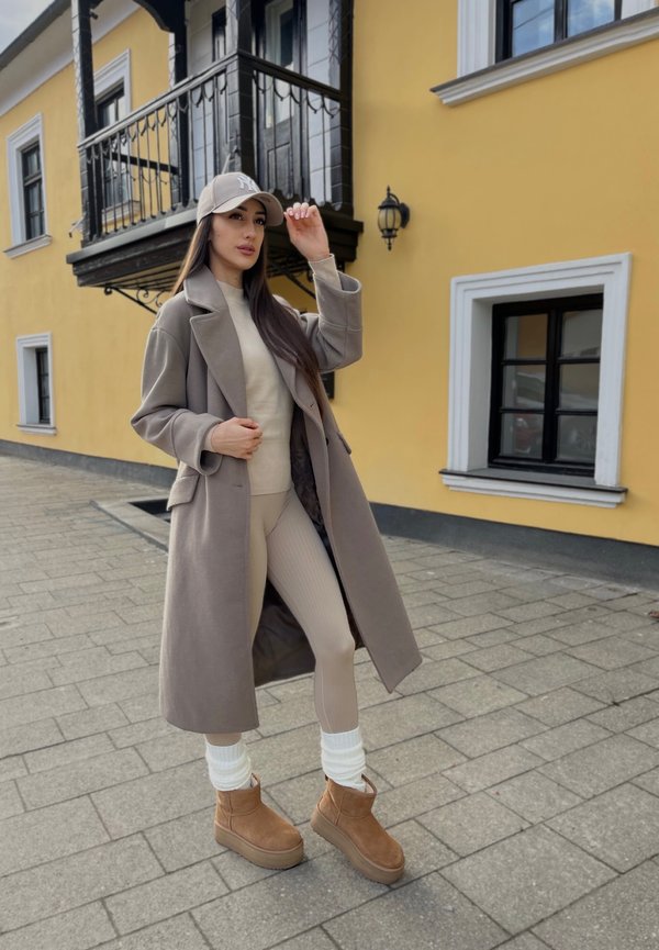 Abrigo beige, suéter crema y leggings de canalé beige claro. Botines marrones con puños de punto blancos. El fondo presenta una pared amarilla y ventanas.