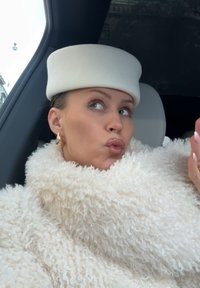 Femme portant un manteau blanc texturé et un chapeau rond assorti à l'intérieur d'une voiture, faisant une expression avec les lèvres pincées tout en regardant sur le côté.