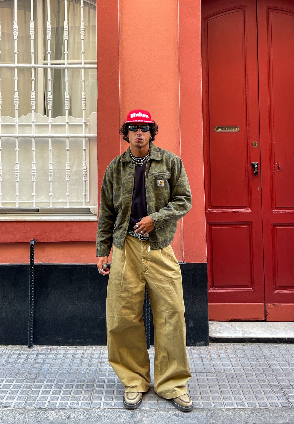 Kamerafarge mønsterjakke, svart t-skjorte, khaki vide bukser, rød caps med hvit tekst, lagdelte halskjeder, solbriller, sko med gummiert såle.